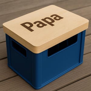May include: A blue storage crate with a light wood lid. The lid is engraved with the word "Papa" in a dark brown font. The crate has rectangular cutouts on the sides for handles. The crate is sitting on a wooden surface.
