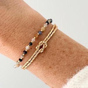 Könnte beinhalten: Zwei Armbänder an einem Handgelenk. Eines ist ein Perlenarmband mit verschiedenen Farben. Das andere ist ein goldfarbenes Armband im Knoten-Design. Die Armbänder werden an einem Handgelenk mit heller Haut getragen.
