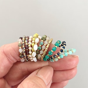 May include: A collection of beaded rings in various colors, including brown, white, green, and blue, with gold accents. The rings are made of small, round beads strung together on a thin band. The rings are displayed on a person's fingers.