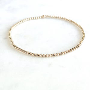 May include: A delicate gold beaded bracelet. The bracelet is composed of small, evenly spaced, round beads. The gold bracelet is displayed on a white marble surface. The bracelet is a simple, elegant piece of jewelry.