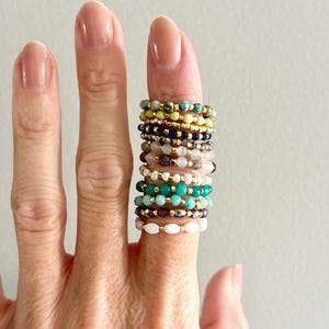 Puede incluir: Una colección de anillos de cuentas en varios colores y diseños. Los anillos están apilados en un dedo, mostrando una variedad de piedras preciosas y tamaños de cuentas. Los colores incluyen verde, dorado, blanco y marrón. Los anillos son delicados y parecen hechos a mano.