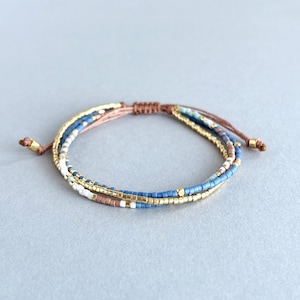 Könnte beinhalten: Ein mehrreihiges Armband mit blauen, weißen und goldenen Perlen. Das Armband ist mit einer braunen Schnur gebunden.