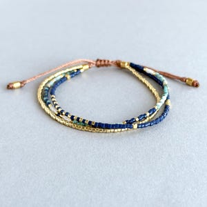 Puede incluir: Un brazalete de múltiples hilos con cuentas azules, verdes y doradas. El brazalete está hecho con un cordón marrón y tiene un cierre de nudo deslizante.
