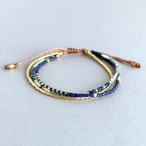 Könnte beinhalten: Ein mehrreihiges Armband mit blauen, goldenen und weißen Perlen. Das Armband ist aus einem braunen Band gefertigt und hat einen verschiebbaren Knoten zum Anpassen der Größe.