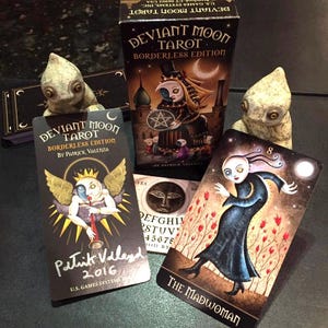 May include: A set of Deviant Moon Tarot cards, a borderless edition by Patrick Valenza. The cards feature dark, whimsical illustrations with a gothic aesthetic. The Madwoman card shows a woman in a black dress with a white face and red flowers in the background. The card is numbered 8.