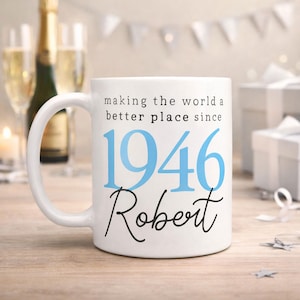 May include: White ceramic mug with the text "making the world a better place since 1946 Robert" in black and blue. The mug has a curved handle and is set against a blurred background with champagne and gifts.