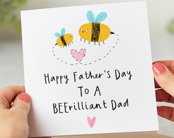 Puede incluir: Una tarjeta blanca con una abeja y una abeja más pequeña con un corazón rosa entre ellas. La tarjeta dice "Happy Father's Day To A BEErilliant Dad" con un corazón rosa en la parte inferior.