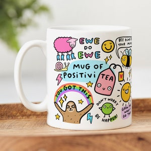May include: White ceramic mug decorated with cheerful cartoon drawings and text. The mug displays a pink sheep, a snail, a sloth under a rainbow, and a tea bag. Text includes "EWE DO EWE", "MUG OF POSITIVI", and "YOU GOT THIS".