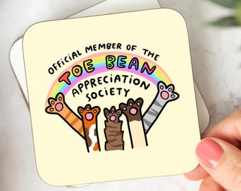 May include: A yellow square coaster with a rainbow and the text "Official Member of the Toe Bean Appreciation Society". Three cat paws with different colored fur are reaching up towards the rainbow.