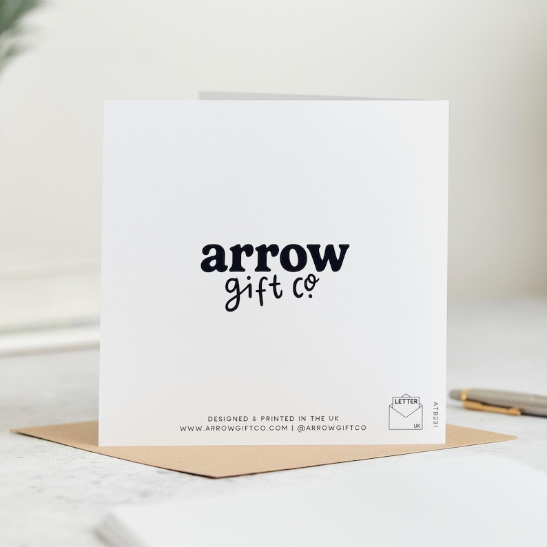 May include: A white greeting card with the text "arrow gift co" in a dark blue font. The card is on a brown envelope. The bottom of the card has the text "DESIGNED & PRINTED IN THE UK" and website and social media information.