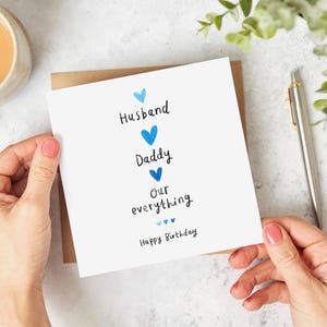Peut inclure: Carte de v&oelig;ux blanche avec l'inscription "Husband, Daddy, Our everything" en &eacute;criture noire, avec des c&oelig;urs bleus. La carte dit aussi "Happy Birthday". La carte est tenue par des mains avec du vernis &agrave; ongles rose.