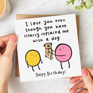 May include: A humorous birthday card featuring a yellow and pink cartoon character with the text "I love you even though you have clearly replaced me with a dog. Happy Birthday!"