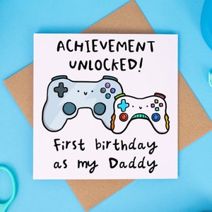 May include: A white card with a cartoon illustration of two video game controllers. The text on the card reads "Achievement Unlocked! First birthday as my Daddy."