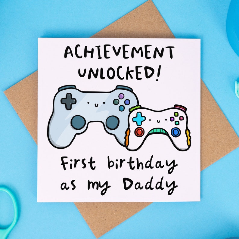 May include: A white card with a cartoon illustration of two video game controllers. The text on the card reads "Achievement Unlocked! First birthday as my Daddy."
