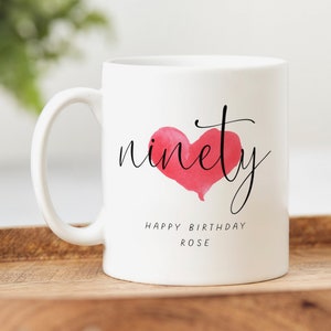Puede incluir: Taza de cerámica blanca con un corazón rosa de acuarela y la palabra "ninety" en escritura negra. El texto "Happy Birthday Rose" está impreso debajo del corazón.