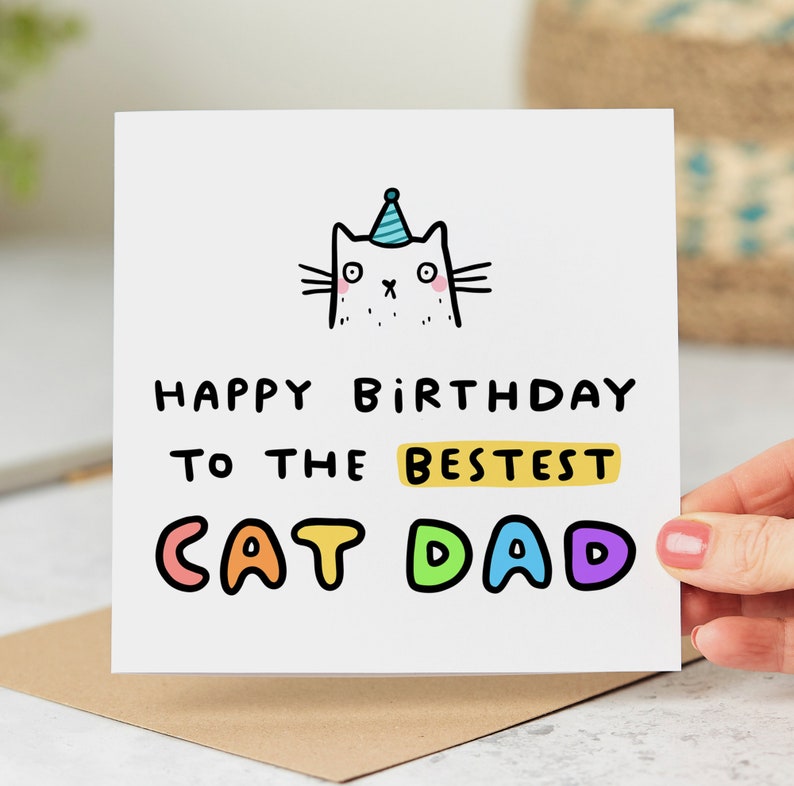 May include: A white card with a cartoon cat wearing a party hat. The card says "Happy Birthday to the Bestest Cat Dad" in colorful letters.