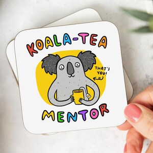 May include: A white coaster with a yellow circle and a cartoon koala holding a cup of tea. The text "KOALA-TEA MENTOR" is written in rainbow colours around the circle. The text "THAT'S YOU!" is written in black next to the koala.