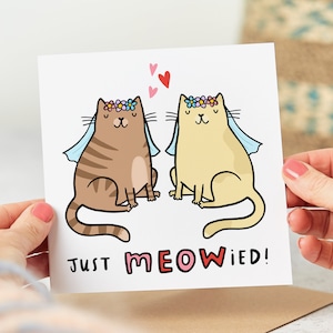 May include: A greeting card featuring a cartoon illustration of two cats wearing flower crowns and veils. The text on the card reads "Just Meowied!"