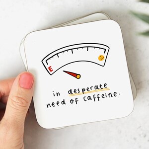 May include: White coaster with a black and white illustration of a fuel gauge with a red needle pointing to "E" and a yellow smiley face. The text reads "in desperate need of caffeine."