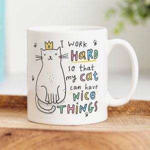 May include: White ceramic mug with a cartoon cat wearing a crown. The mug has the text "I work hard so that my cat can have nice things" in colorful letters.