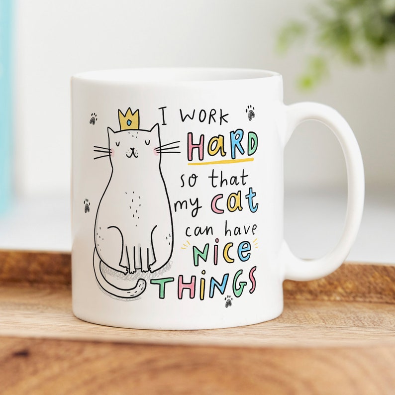 May include: White ceramic mug with a cartoon cat wearing a crown. The mug has the text "I work hard so that my cat can have nice things" in colorful letters.