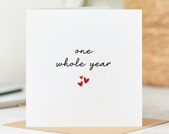 Può includere: Un biglietto bianco con testo nero che recita "one whole year" e tre piccoli cuori rossi.