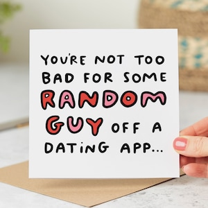 May include: A white card with black text that reads "You're not too bad for some random guy off a dating app..." The words "random guy" are in pink and red.