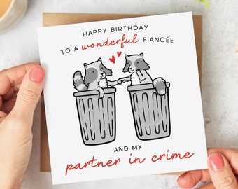 Puede incluir: Tarjeta de felicitación blanca con sobre marrón. La tarjeta presenta una ilustración de dibujos animados de dos mapaches tomados de la mano en botes de basura, con el texto "Happy Birthday to a wonderful fiancée and my partner in crime."