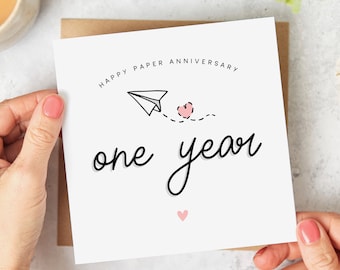 Può includere: Un biglietto bianco con il testo "Happy Paper Anniversary one year" in corsivo nero. Un aeroplano di carta con un cuore rosa è disegnato sopra il testo. Un piccolo cuore rosa si trova in fondo al biglietto.