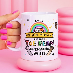 May include: White ceramic mug featuring a rainbow, a cat, and the text "Official Member of the Toe Bean Appreciation Society". The name "Amelia" is at the top of the mug.