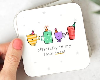May include: A stack of white square coasters with a whimsical design. Each coaster features a cartoon teacup with a party hat. The text "officially in my four-teas!" is printed below the teacups. Small star shapes are scattered around the design.