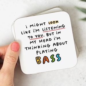 May include: White coaster with black text that reads "I might look like I'm listening to you. But in my head I'm thinking about playing bass". The word "bass" is written in colourful letters.