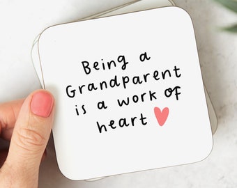 May include: White coaster with black text that reads "Being a Grandparent is a work of heart" with a pink heart.
