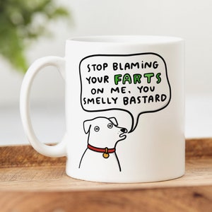 May include: White ceramic mug featuring a cartoon dog with a red collar and gold tag. The speech bubble says, "STOP BLAMING YOUR FARTS ON ME, YOU SMELLY BASTARD." The word "FARTS" is in green.