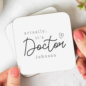 May include: White square coaster with the text "actually, it's Doctor Johnson" in black script and a small heart symbol. The coaster is part of a stack of coasters.