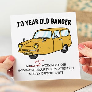 May include: A funny birthday card featuring a cartoon illustration of a yellow car with the text "70 YEAR OLD BANGER" and "B4NG3R" on the number plate. The card also includes the text "decent, IN PERFECT WORKING ORDER, BODYWORK REQUIRES SOME ATTENTION, MOSTLY ORIGINAL PARTS."