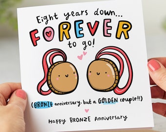 May include: A square white card with the text "Eight years down... FOREVER to go!" and "Happy BRONZE Anniversary." Features two cartoon medals with pink ribbons. The card is held by two hands.
