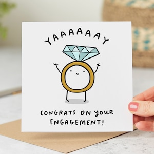 May include: A white card with a cartoon drawing of a ring with a diamond on top. The ring has a happy face and arms raised in the air. The text "YAAAAAY" is above the ring and "CONGRATS ON YOUR ENGAGEMENT!" is below the ring.