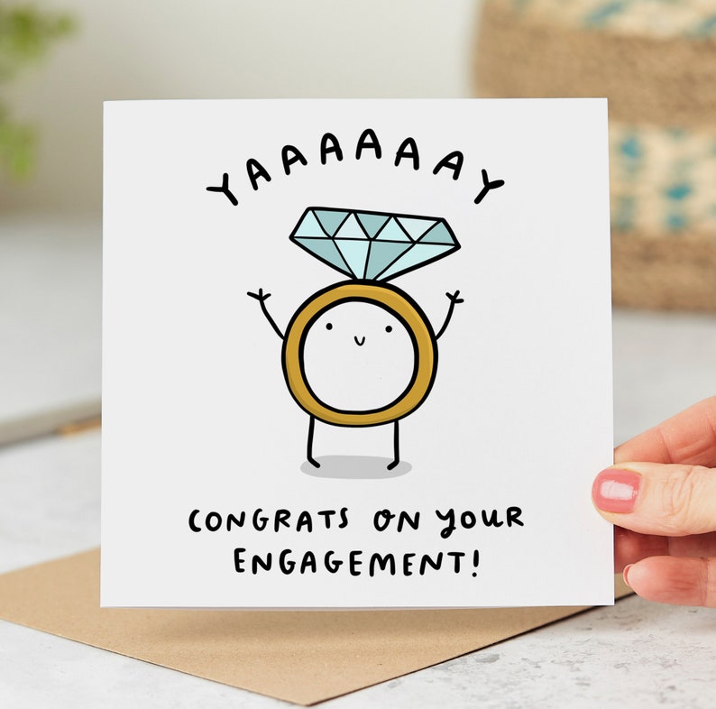 May include: A white card with a cartoon drawing of a ring with a diamond on top. The ring has a happy face and arms raised in the air. The text "YAAAAAY" is above the ring and "CONGRATS ON YOUR ENGAGEMENT!" is below the ring.