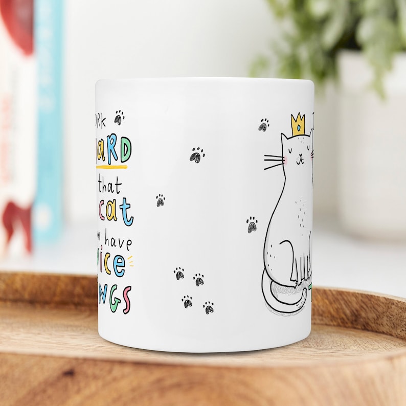 May include: White ceramic mug with a black and white illustration of a cat wearing a gold crown. The text on the mug reads "Work hard that cat can have nice things".