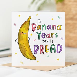 May include: A square greeting card featuring a cartoon banana and the phrase "In Banana Years You're BREAD". The banana is yellow with a smiling face and arms. The text is in colourful bubble letters, with confetti and stars.