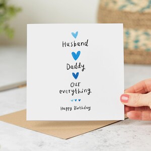 Peut inclure: Carte de v&oelig;ux blanche avec des c&oelig;urs bleus et les mots "Husband, Daddy, Our everything, Happy Birthday." La carte est tenue au-dessus d'une enveloppe marron.