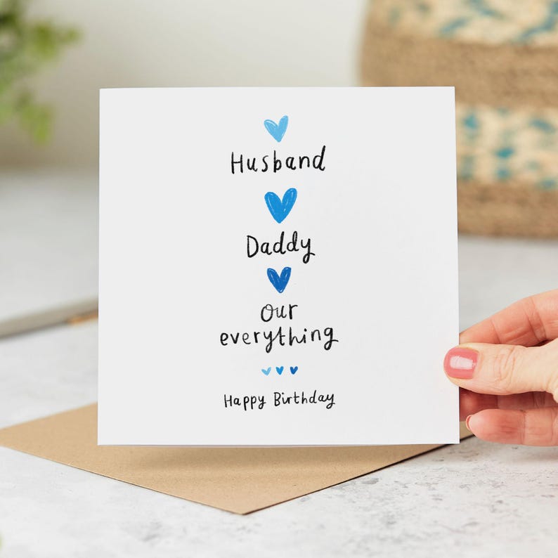 Peut inclure: Carte de v&oelig;ux blanche avec des c&oelig;urs bleus et les mots "Husband, Daddy, Our everything, Happy Birthday." La carte est tenue au-dessus d'une enveloppe marron.