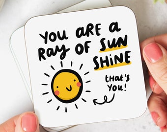 May include: White coaster with black text that reads "You are a ray of sunshine that's you!" with a yellow sun with a smiling face and red cheeks.