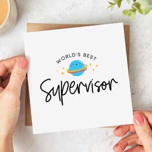May include: A white card with the text "World's Best Supervisor" in black lettering. A blue planet with a yellow ring and a smiling face is above the text.