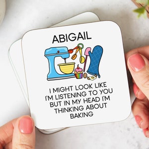 May include: White coaster with the name Abigail and a cartoon illustration of a stand mixer, cupcake, and other baking supplies. The text reads "I might look like I'm listening to you but in my head I'm thinking about baking."