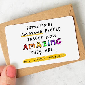 May include: A white card with the text "Sometimes amazing people forget how amazing they are... this is your reminder!" The text is written in black marker with a yellow underline under "amazing people" and a yellow underline under "this is your reminder!" The word "amazing" is written in rainbow colours.