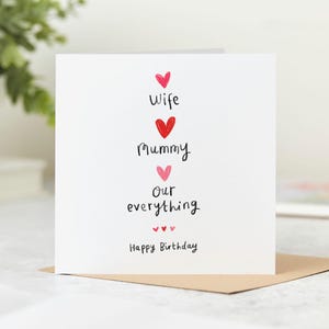 May include: A white greeting card with the words "Wife, Mummy, Our everything" in black script, with red and pink heart accents. The words "Happy Birthday" are at the bottom. The card is on a brown envelope.