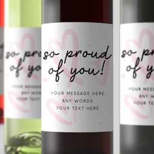 May include: A white wine bottle label with pink hearts and the text "so proud of you!"  The label also includes the text "YOUR MESSAGE HERE ANY WORDS YOUR TEXT HERE".