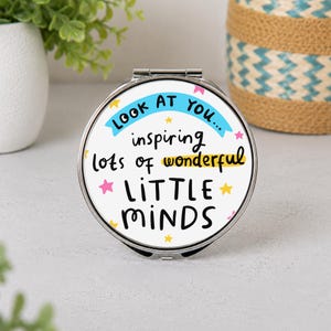 May include: A round silver compact mirror featuring a white surface with the text "LOOK AT YOU... inspiring lots of wonderful LITTLE MINDS" in black and blue lettering, accented by small pink and yellow stars scattered across the design.